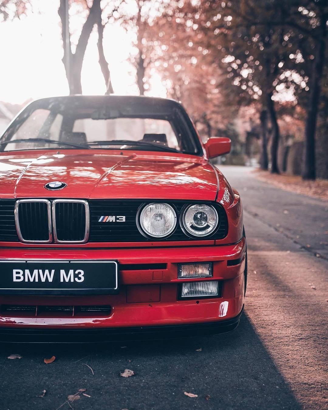 Red BMW E30 M3 sportscar parked on a city street with the iconic BMW logo on the grill and the M3 badge on the back