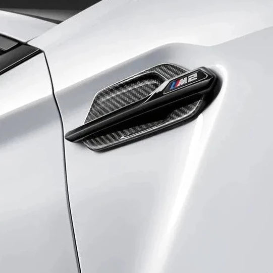 Close-up view of a BMW M2's side fender vent made of sleek carbon fiber material. The vent features a stylish, modern design with a horizontal strip of small perforations for improved airflow. The carbon fiber texture adds a high-performance, racing-inspired look to the car's exterior.